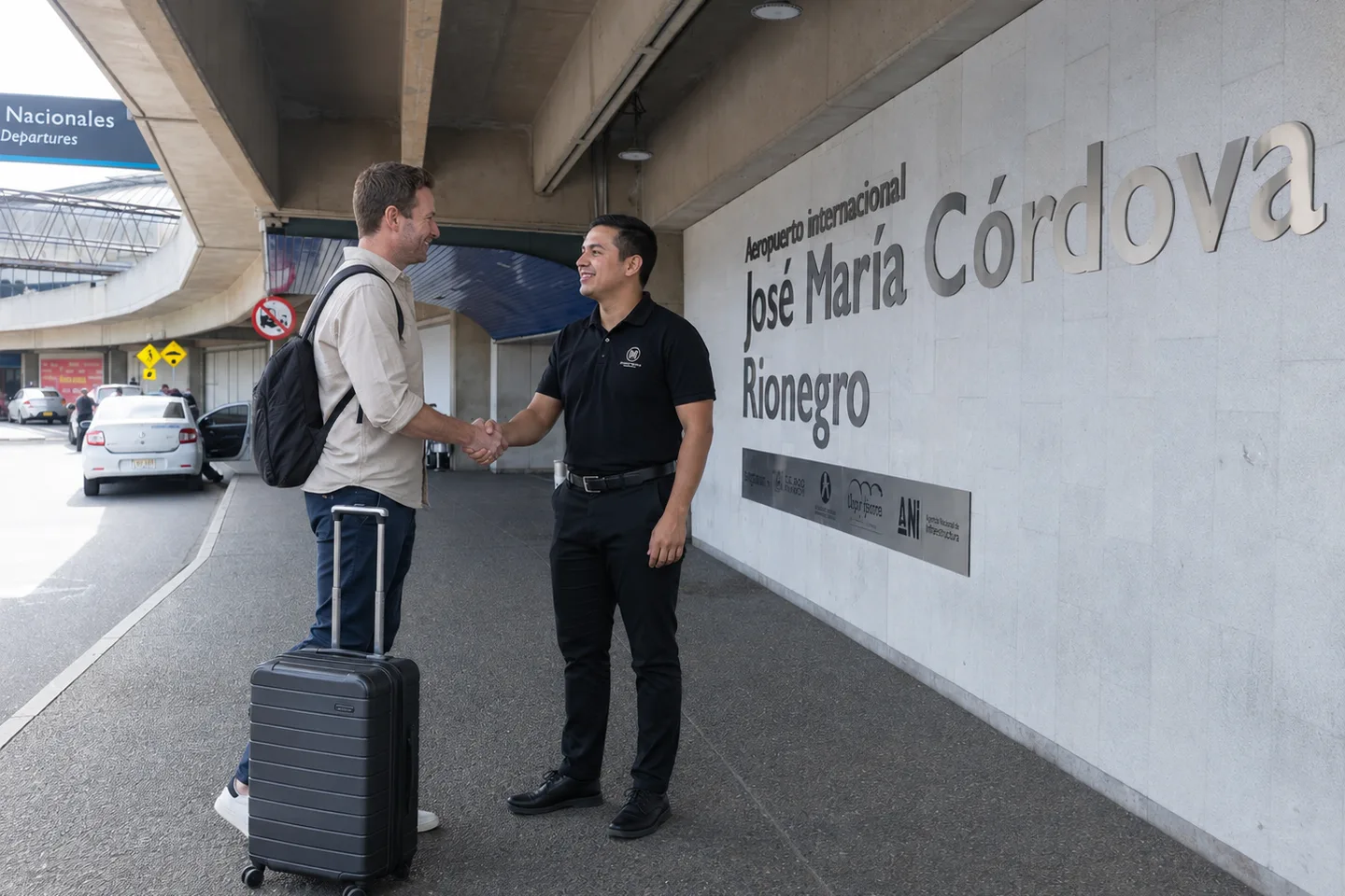 Un asesor de Majestic con camisa negra saluda de la mano a un viajero con maleta en la entrada del Aeropuerto Internacional José María Córdova, en Rionegro.